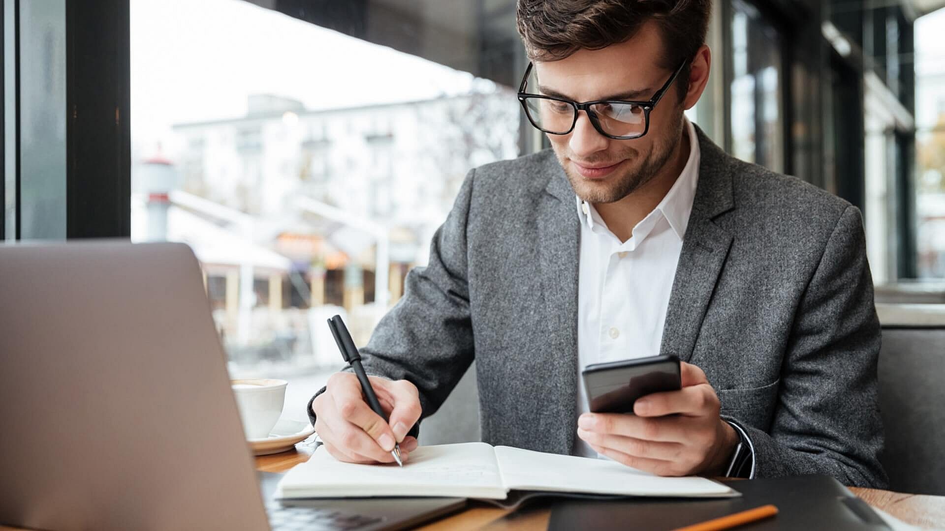 Smiling businessman eyeglasses sitting by table cafe with laptop computer while using smartphone writing something 1 Smiling businessman eyeglasses sitting by table cafe with laptop computer while using smartphone writing something 1
